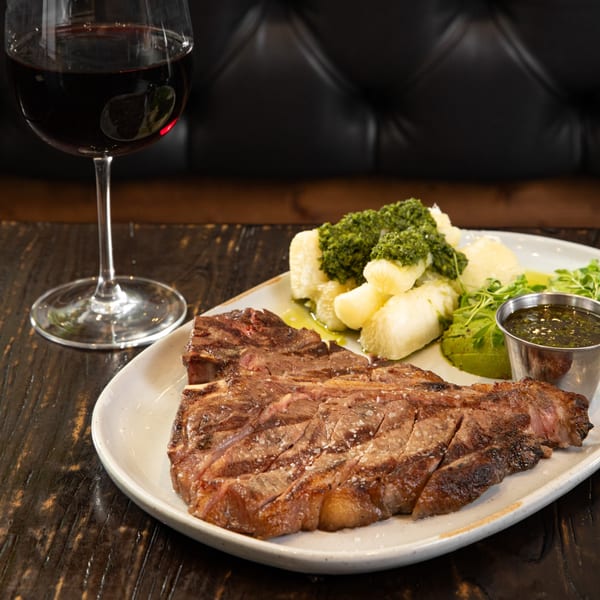 Grilled steak with yucca, green sauce, avocado, and a glass of red wine on a wooden table.