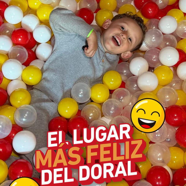 A happy child smiles while lying in a ball pit filled with red, yellow, and white balls.
