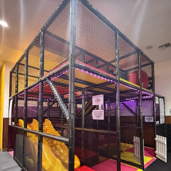A large, multi-level indoor playground with black netting, yellow slides, and purple accent lighting.