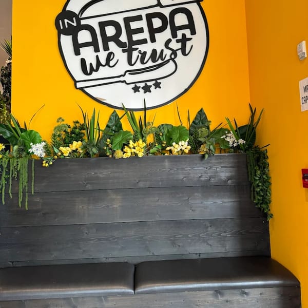 A black wooden bench with plants and a "In Arepa We Trust" sign on a bright yellow wall.