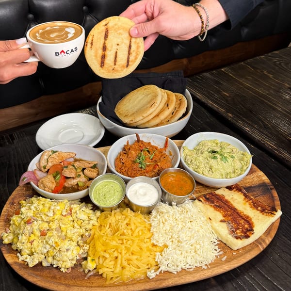 Hands holding a Bocas Grill latte and an arepa, with a large Latin-American breakfast platter.