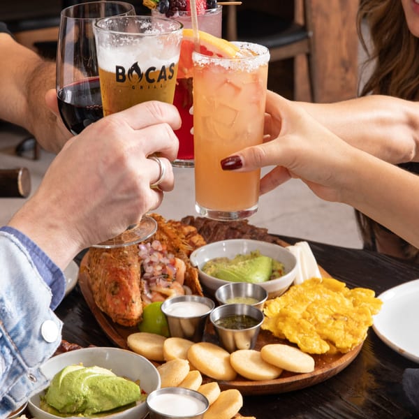 Four people toasting drinks, including a Bocas Grill beer, over a large Latin-American food platter.