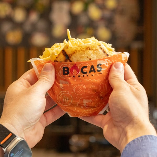 Hands holding a Bocas Grill arepa overflowing with shredded cheese and french fries.
