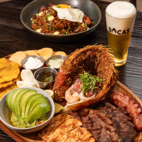 Bocas Grill's Latin-American feast: fried fish, grilled meats, avocado, rice, and beer on a wooden table.