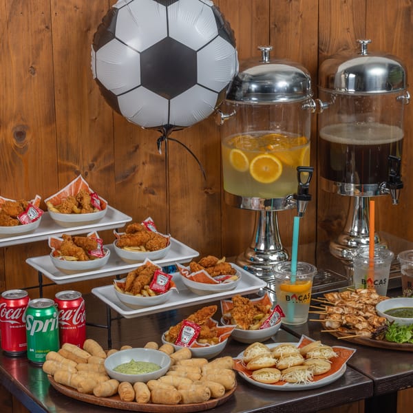 A soccer-themed party spread with Bocas Grill chicken tenders, tequeños, arepas, chicken skewers, and drinks.
