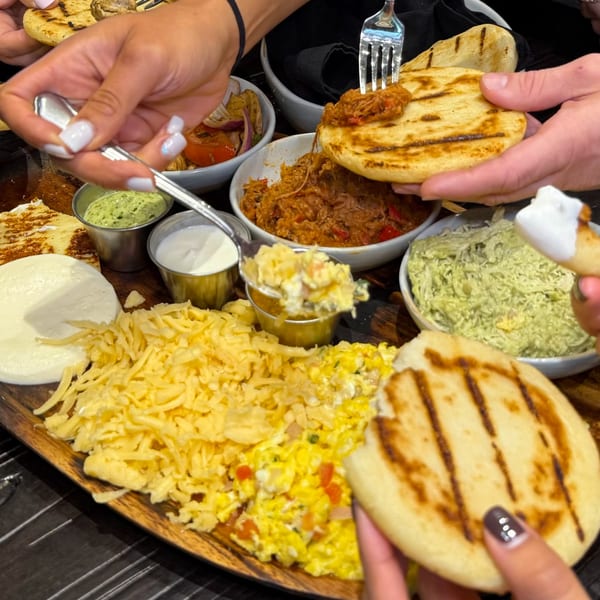Hands sharing a large platter of Latin-American food, including arepas, shredded meat, and various toppings.
