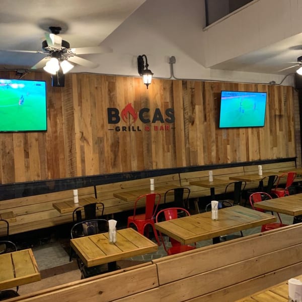 Interior of Bocas Grill & Bar, featuring wooden tables, red and black chairs, and TVs on a wood-paneled wall.