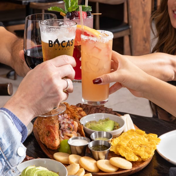 Four people toasting drinks, including a Bocas Grill beer, over a large Latin-American food platter.