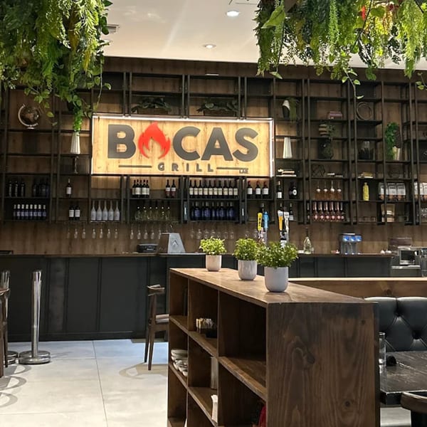 The interior of Bocas Grill features a large wooden sign, a bar with wine bottles, and hanging plants.
