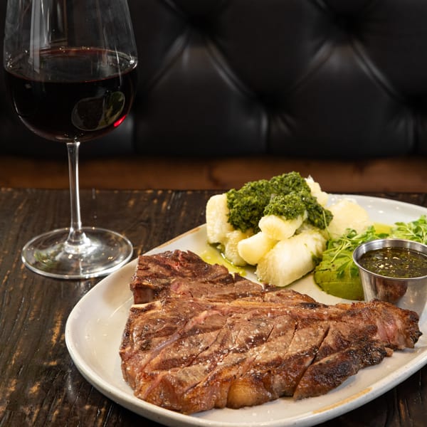 Grilled steak with yucca, chimichurri, avocado, and a glass of red wine on a dark table.
