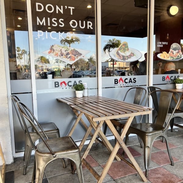 Outdoor seating area at Bocas Grill with a wooden table, metal chairs, and window decals of Latin-American food.