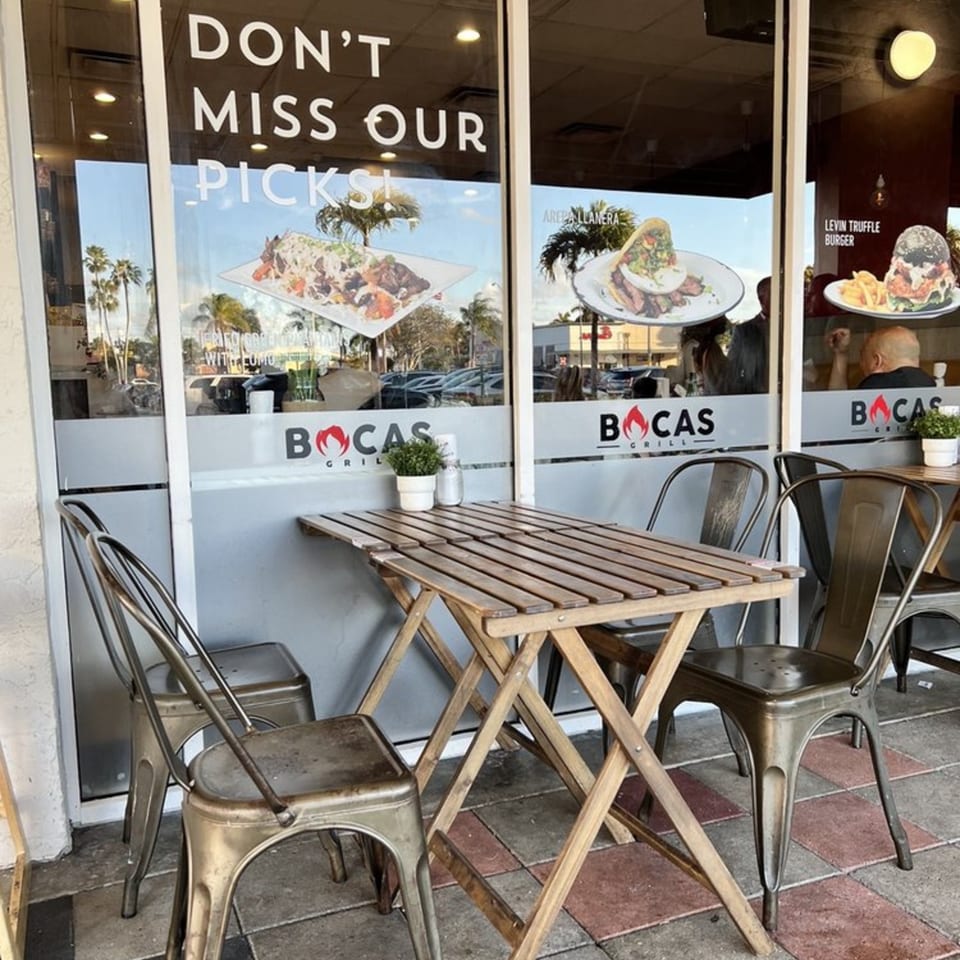 Outdoor seating area at Bocas Grill with a wooden table, metal chairs, and window decals of Latin-American food.
