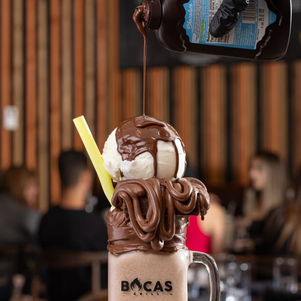 A hand pours chocolate syrup onto a Bocas Grill milkshake topped with ice cream and brownie.